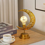 A gold crescent-moon table lamp with a glowing light bulb in the center, sitting on a wooden desk beside stacked books and a white teacup.