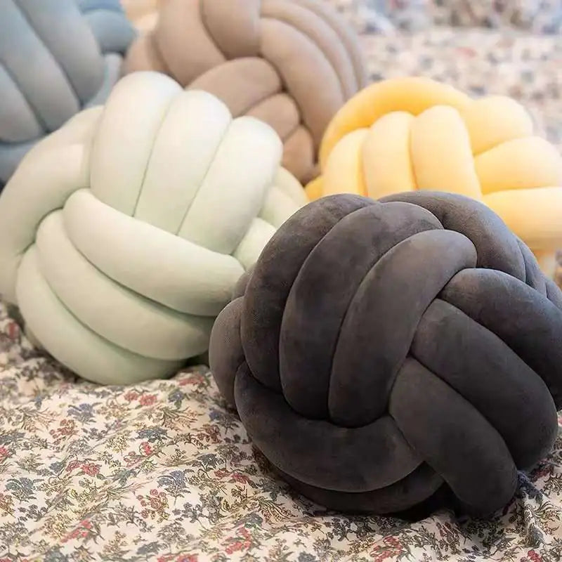 Several soft knotted ball pillows in pastel and neutral colors, including mint green, gray, beige, blue, and yellow, arranged on a floral-patterned bedspread.