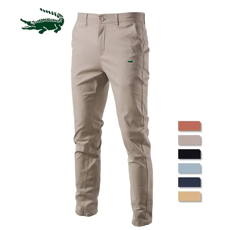 A pair of slim-fit khaki pants with a button and zipper closure, featuring a small green crocodile logo on the upper left thigh. The pants have belt loops and side pockets, with a slightly wrinkled appearance. A larger green crocodile logo is displayed in the top left corner of the image.