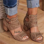 A close-up view of a person wearing stylish beige cut-out heeled sandals with ankle straps and decorative perforations, paired with cuffed blue denim jeans on a wooden floor.