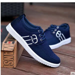 A pair of navy blue casual canvas sneakers with white rubber soles and lace-up fronts, displayed on a wooden floor. One shoe is propped against a shoebox, while the other sits in the background, and both feature white side stripe detailing with “SPORT” printed on the outer side.
