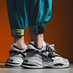 White chunky sneakers with black strap accents, thick layered soles, and lace-up design, shown worn with teal jogger pants against an orange background.