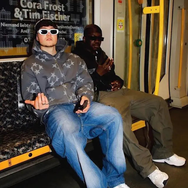 Two people wearing sunglasses sit on a subway/train seat; one in a gray star-pattern puffer jacket and jeans holds a phone, while the other wears dark clothing and olive pants.