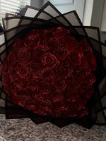 A bouquet of deep red glittery roses wrapped in layered black translucent paper, displayed on a countertop.