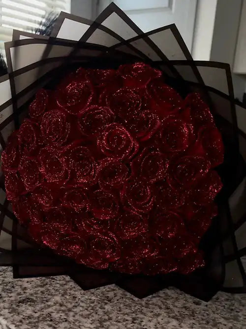 A bouquet of deep red glittery roses wrapped in layered black translucent paper, displayed on a countertop.