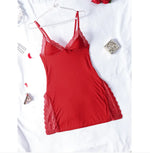 Woman wearing a black satin and lace lingerie nightdress with spaghetti straps, deep V-neckline, and lace trim along the neckline and high side slits. The outfit features a small bow detail at the center and is styled in a softly lit bedroom setting.