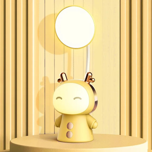 A cute yellow character-shaped desk lamp with a glowing oval face, closed smiling eyes, and small antenna-like details on top. A round light panel extends upward from the back on a slim white stem, while metallic gold accents add a polished look. The lamp is displayed on a matching round platform against a warm yellow striped background.