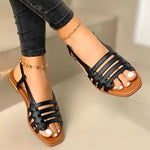 A pair of black strappy flat sandals worn with skinny black pants, featuring open toes, slingback straps, and tan footbeds. The model is also wearing gold ankle bracelets, and the sandals are shown on a light beige indoor surface.