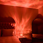 A moody bedroom scene lit in deep red, with a small lamp or projector on a nightstand casting swirling, flame-like light patterns across the wall and ceiling. A tufted headboard and pillows are visible on the left, and an arched floor mirror and chair sit on the right.