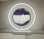 A tabletop circular LED ring lamp with a bright white light, sitting on a rectangular base. In the center of the ring is a round sand art frame showing purple sand at the top and bottom with white sand in the middle, forming layered “mountain” shapes as the sand trickles downward. The photo is taken against a plain wall in a simple indoor setting.