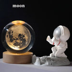 Crystal ball night light with a glowing moon design on a wooden base beside a small astronaut figurine kneeling on a moon-like surface, with the word “moon” at the top.