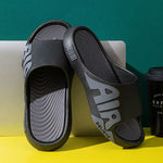 A pair of futuristic open-toe slide sandals with a sleek, curved design and large side cutouts, displayed on a minimalist gray and white background. An inset image shows a beige version of the sandals worn with black socks.