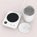 White electric cup warmer with a black heating plate and digital display, shown beside a white mug and a matching lid on a light background.