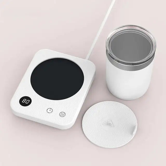 White electric cup warmer with a black heating plate and digital display, shown beside a white mug and a matching lid on a light background.