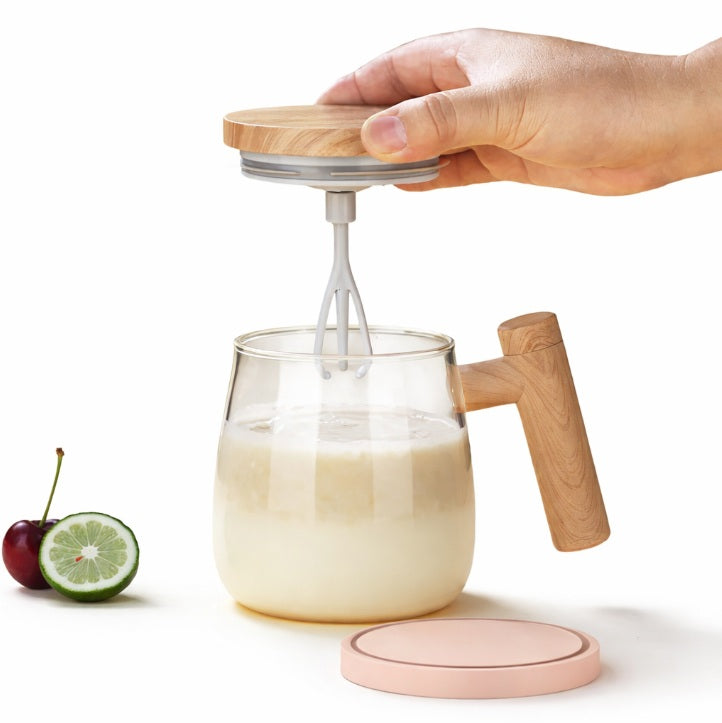 Hand using a hand mixer to blend liquid in a glass with wooden handle and lid on a white background.