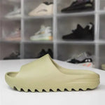 A beige Yeezy Slide with a thick, ridged sole displayed on a white surface, with a blurred background of various sneakers in transparent shoe storage boxes.