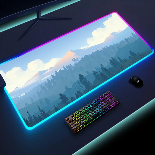 Large gaming mouse pad with a mountain-and-forest landscape design and glowing RGB light strip around the edges, shown on a desk with a rainbow-backlit keyboard and a mouse.