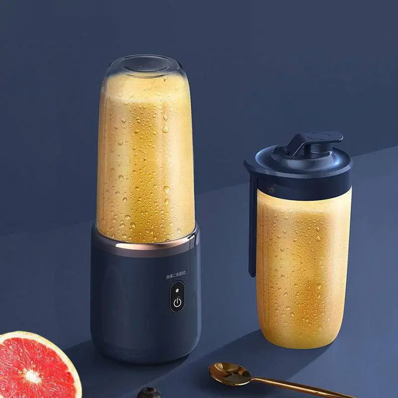 Portable blender with a dark base and an upside-down blending cup filled with orange-colored smoothie, shown beside a matching to-go cup with lid and handle on a dark surface, with a grapefruit slice and spoon nearby.