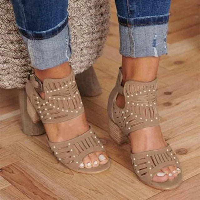 A close-up view of a person wearing stylish beige cut-out heeled sandals with ankle straps and decorative perforations, paired with cuffed blue denim jeans on a wooden floor.