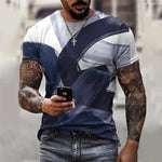 A man wearing a fitted short-sleeve T-shirt with a bold abstract navy-and-white geometric print, paired with blue jeans. He has tattooed arms, a cross necklace, and is holding a smartphone while standing outdoors in a blurred street setting.