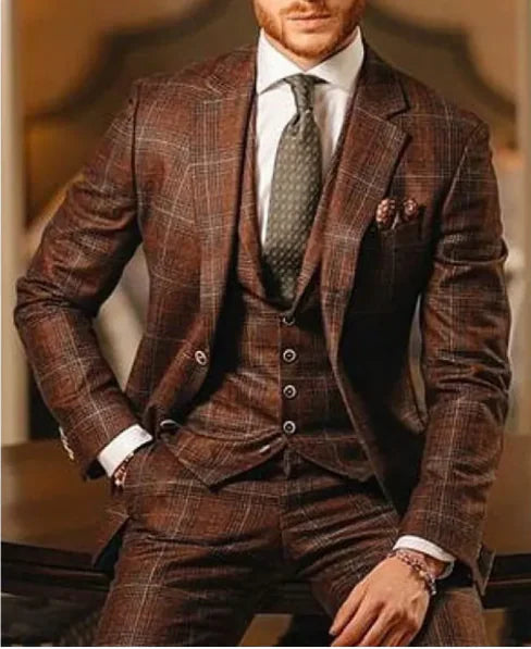 A sophisticated man dressed in a three-piece brown plaid suit with a subtle checkered pattern. The ensemble includes a tailored blazer, a matching vest, and trousers, paired with a crisp white dress shirt and a dark green textured tie. A patterned pocket square adds a refined touch. He accessorizes with a bracelet on his wrist. The warm, elegant background complements the rich tones of his suit, enhancing the overall luxurious aesthetic.