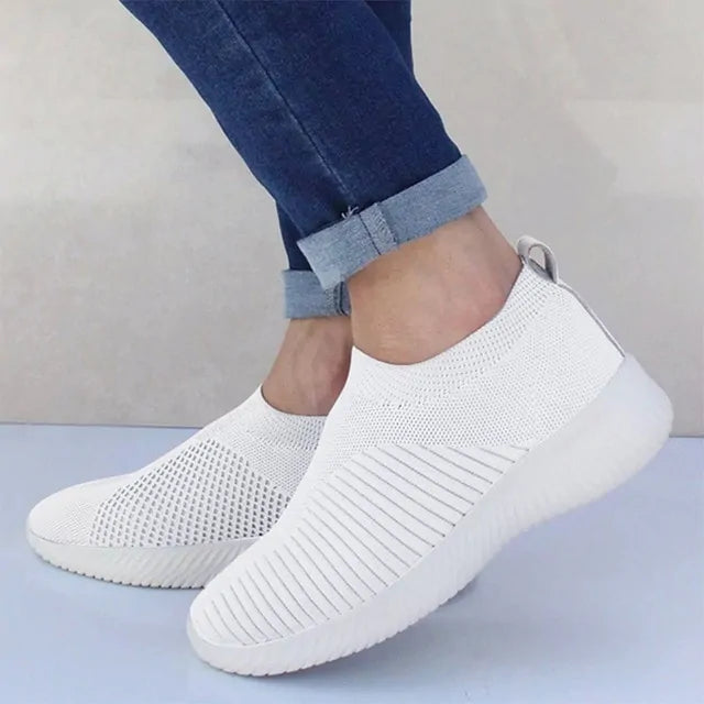 White slip-on knit sneakers worn with rolled-up blue jeans.