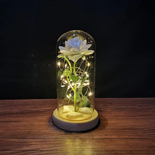 Artificial white rose inside a glass dome, wrapped with warm string lights on a wooden base, displayed on a table against a dark background.