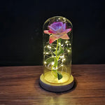 Blue rose with gold stem and leaves inside a glass dome, wrapped with warm white fairy lights, sitting on a wooden base against a dark background.