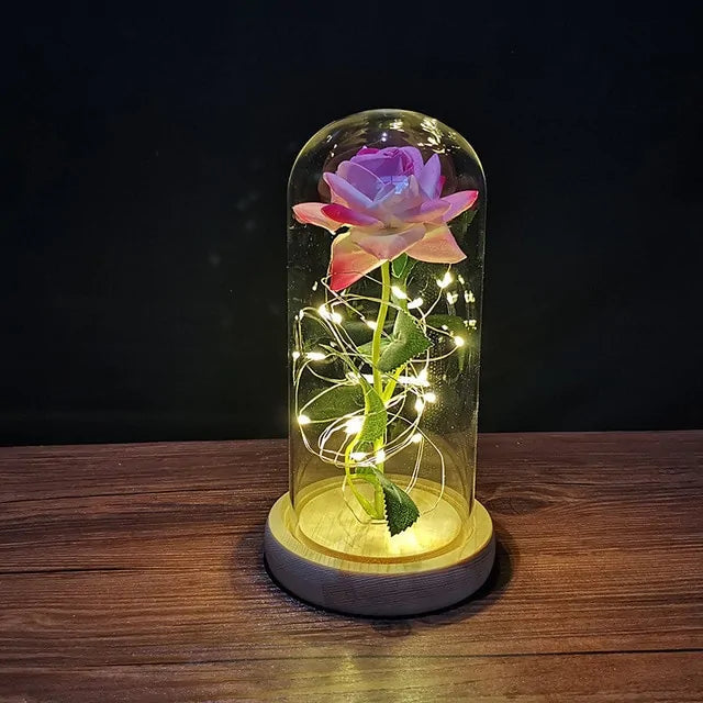 Blue rose with gold stem and leaves inside a glass dome, wrapped with warm white fairy lights, sitting on a wooden base against a dark background.