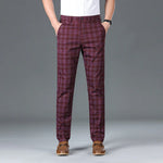 A man wearing slim-fit, beige plaid trousers with a black checkered pattern. The trousers have a classic button and zipper closure, belt loops, and side pockets. He pairs them with a white button-up shirt tucked in, brown leather loafers with a decorative metal accent, and a black wristwatch. The background is a simple gray backdrop, emphasizing the stylish and sophisticated outfit.