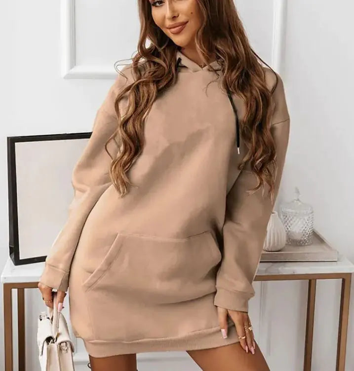 Woman wearing a long-sleeve, oversized maroon hoodie dress with a front kangaroo pocket and gold drawstrings. She is holding a light-colored handbag and posing indoors with a soft smile. The outfit has a casual, cozy look perfect for lounging or casual outings.