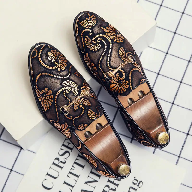 A pair of elegant burgundy leather loafers with a glossy finish. The shoes feature gold-toned metal horsebit detailing across the uppers and are displayed with wooden shoe trees inserted. They have a sleek, elongated silhouette with subtle stitching along the toe cap. The loafers are placed on a dark surface, partially framed by a wooden picture frame, emphasizing their classic and sophisticated style.