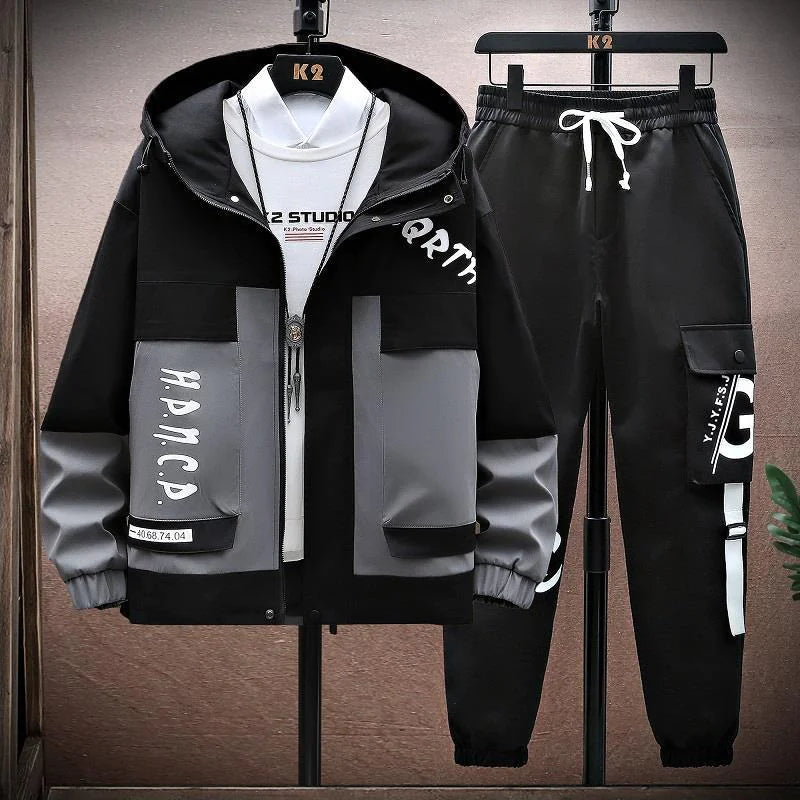 A stylish men's streetwear tracksuit set displayed on hangers. The set includes a black and teal hooded jacket with a bold, modern design, featuring large pockets, graphic text details, and a front zipper closure. The matching black jogger pants have a relaxed fit, cargo-style pockets, and white graphic text on the side, along with a drawstring waistband for adjustability. The outfit is paired with a white t-shirt and a pendant necklace, giving it a trendy, urban aesthetic.