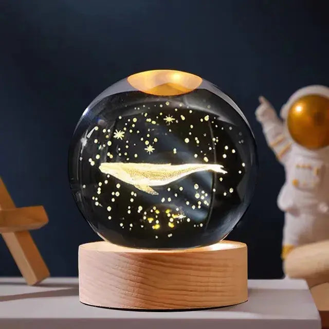 A glowing glass crystal ball lamp on a wooden base, displaying an illuminated whale surrounded by star-like dots, with a small astronaut figurine in the background.