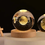 Close-up of a glowing glass crystal ball on a round wooden base, etched with a golden ringed planet and tiny stars inside; warm ambient lights reflect on the sphere against a dark background, with another similar sphere partially visible to the right.