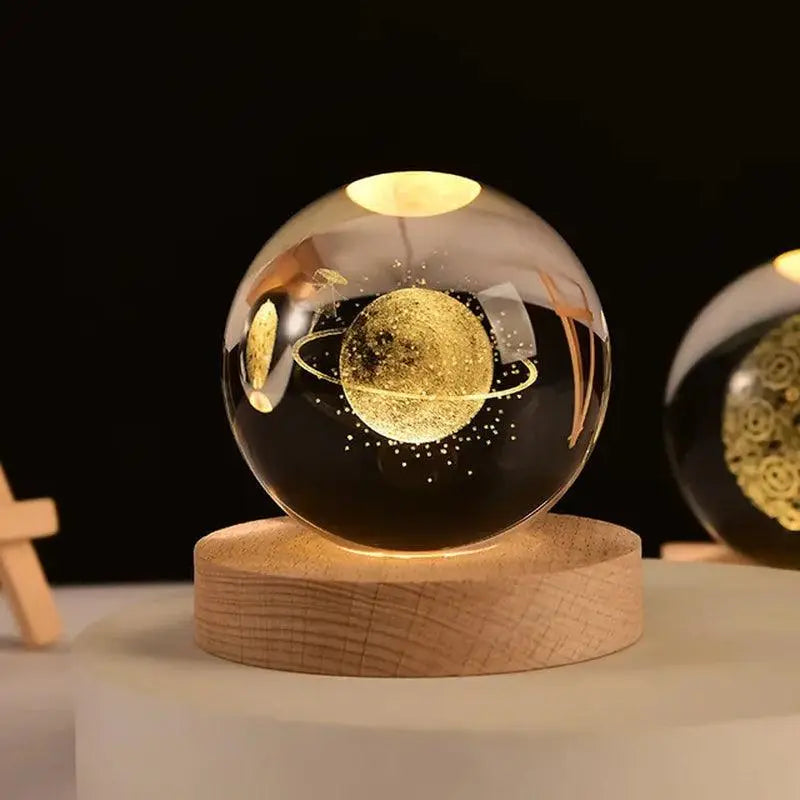 Close-up of a glowing glass crystal ball on a round wooden base, etched with a golden ringed planet and tiny stars inside; warm ambient lights reflect on the sphere against a dark background, with another similar sphere partially visible to the right.