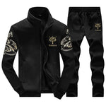 A studio product image of a black two-piece tracksuit set: a zip-up jacket with a stand collar and side pockets, and matching jogger pants with a drawstring waist. Both pieces feature a small gold crest-style logo and the word “CLASSISAL,” plus white ornamental/scroll patterns on the sleeves and upper legs, shown on a white background.