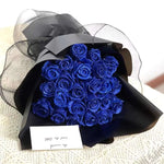 A bouquet of deep blue glitter roses wrapped in matte black paper and black sheer mesh, arranged in a dramatic gift-style presentation. It is shown with a small note card, giving the bouquet a romantic and elegant look.