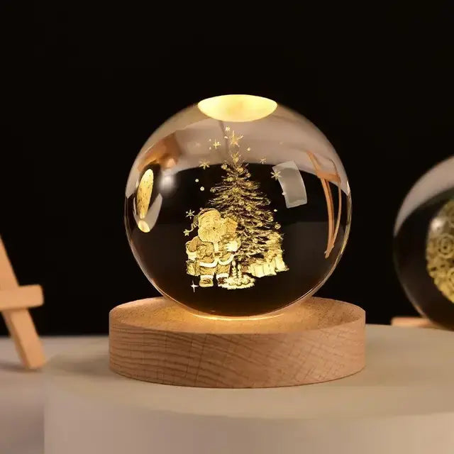 A warm-lit crystal ball night light sitting on a round wooden base against a dark background. Inside the clear glass sphere is a laser-etched Christmas scene—a tree with small star/snowflake details and Santa with a child (or a small figure beside Santa). The glow is golden/amber, making the etched design stand out.