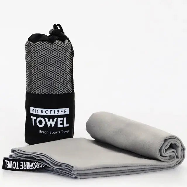 A microfiber towel in a black mesh carrying bag with the label "MICROFIBER TOWEL" on it. The towel is light grey, unfolded and lying flat, showing a soft, smooth texture.