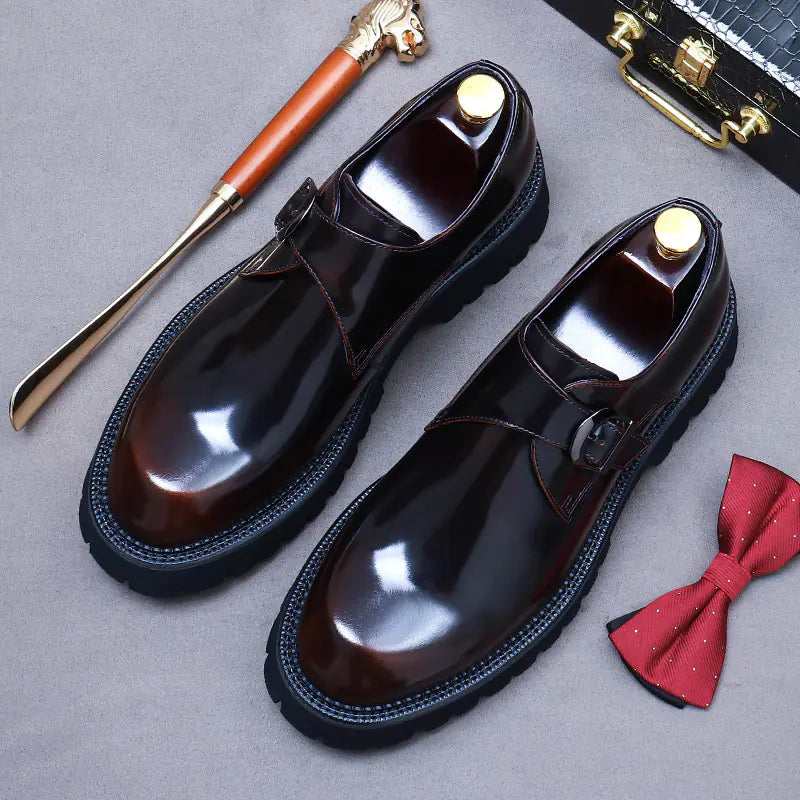A pair of elegant burgundy leather loafers with a glossy finish. The shoes feature gold-toned metal horsebit detailing across the uppers and are displayed with wooden shoe trees inserted. They have a sleek, elongated silhouette with subtle stitching along the toe cap. The loafers are placed on a dark surface, partially framed by a wooden picture frame, emphasizing their classic and sophisticated style.