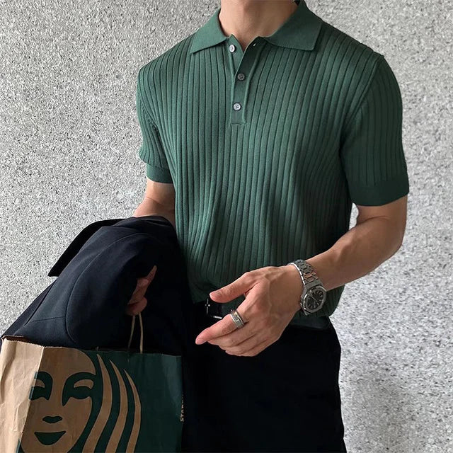 A stylish man wearing a fitted, ribbed green polo shirt with short sleeves and a buttoned collar. He is accessorized with a silver wristwatch and multiple rings. In one hand, he holds a black blazer, while in the other, he carries a Starbucks paper bag. The background features a textured, neutral-coloured wall.