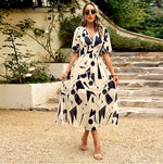 A woman standing outdoors wearing a vibrant floral print midi dress in shades of pink, red, and white. The dress features a V-neckline, puffed elbow-length sleeves
