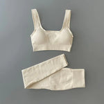 Black ribbed sports bra and matching high-waisted leggings set laid flat on a neutral gray background. The outfit is designed for activewear or athleisure, featuring a scoop neckline and wide straps on the bra.