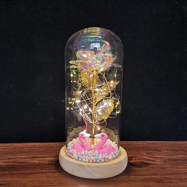Decorative LED lamp under a clear glass dome on a wooden base, featuring a shimmering rainbow-like rose wrapped in warm fairy lights, with pastel pebble accents and two small pink flamingo figures at the bottom against a dark background.
