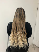 Back view of a person wearing long ombré box braids that transition from dark roots to honey blonde, ending in loose curly blonde tips, standing indoors in a black outfit near a white door.
