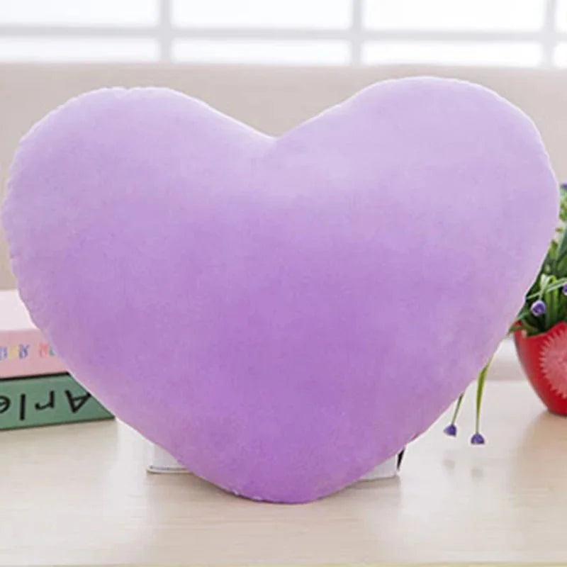 Soft plush heart-shaped pillow in light purple, sitting on a tabletop with a blurred background.
