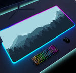 RGB-edged extended gaming mouse pad with a minimalist mountain-and-forest silhouette design, placed on a dark desk next to a rainbow-backlit keyboard and a black mouse.