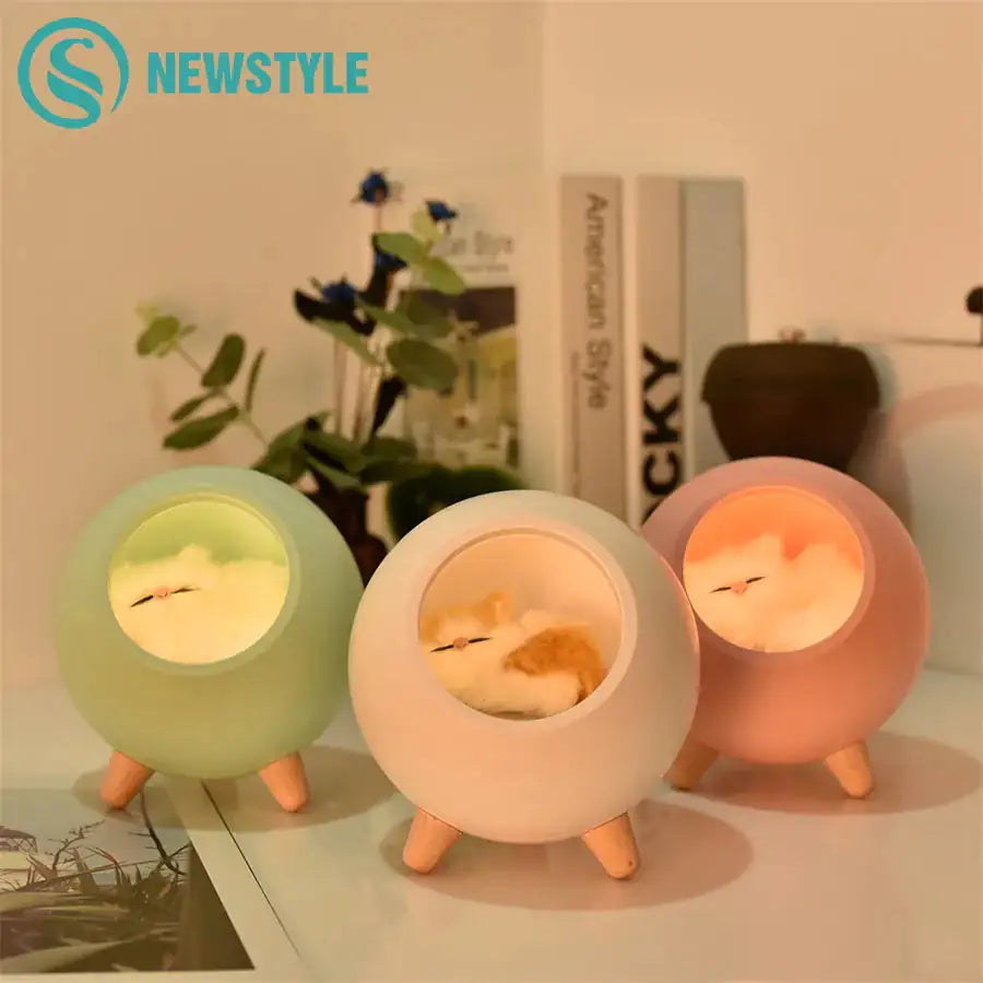 Three round bedside night lights with short wooden legs, glowing in green, white, and pink, each featuring a sleeping bird design inside the center window.