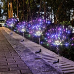 Two dandelion-style outdoor stake lights glowing in a grassy yard at night, with bright white “spark” branches and trees silhouetted in the background.